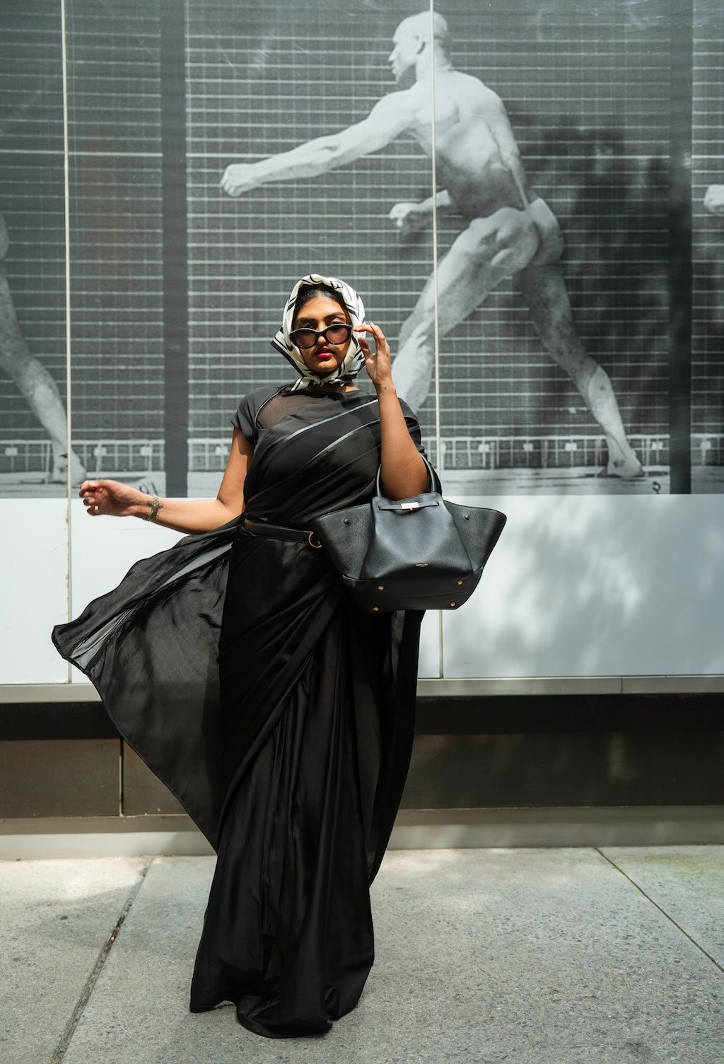Woman in Rebel Roots Capsule Black Saree and scarf holding a gray bag, standing in front of a glass wall with a sculpture.