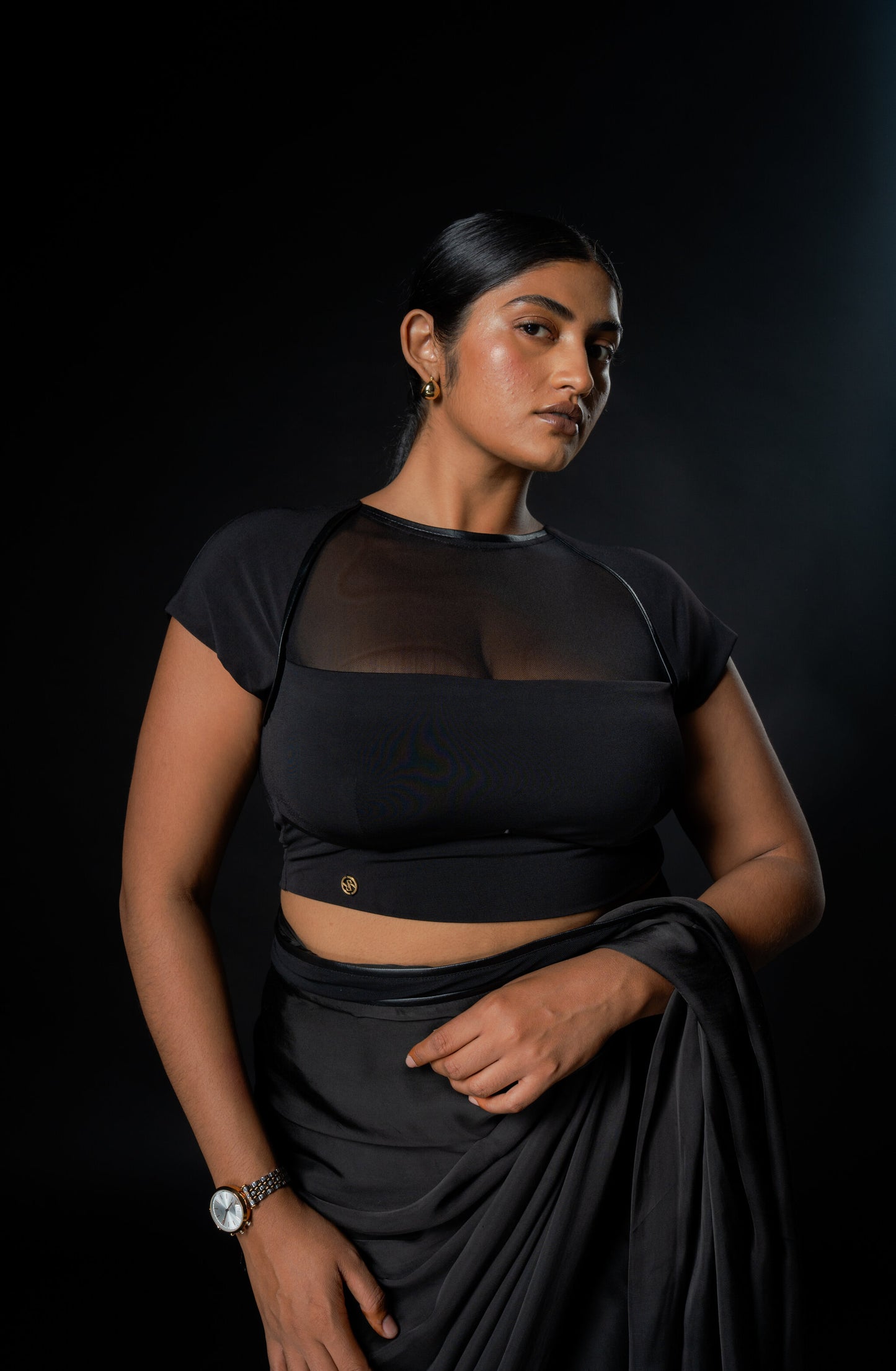 Woman wearing a black saree with a sheer blouse against a dark background