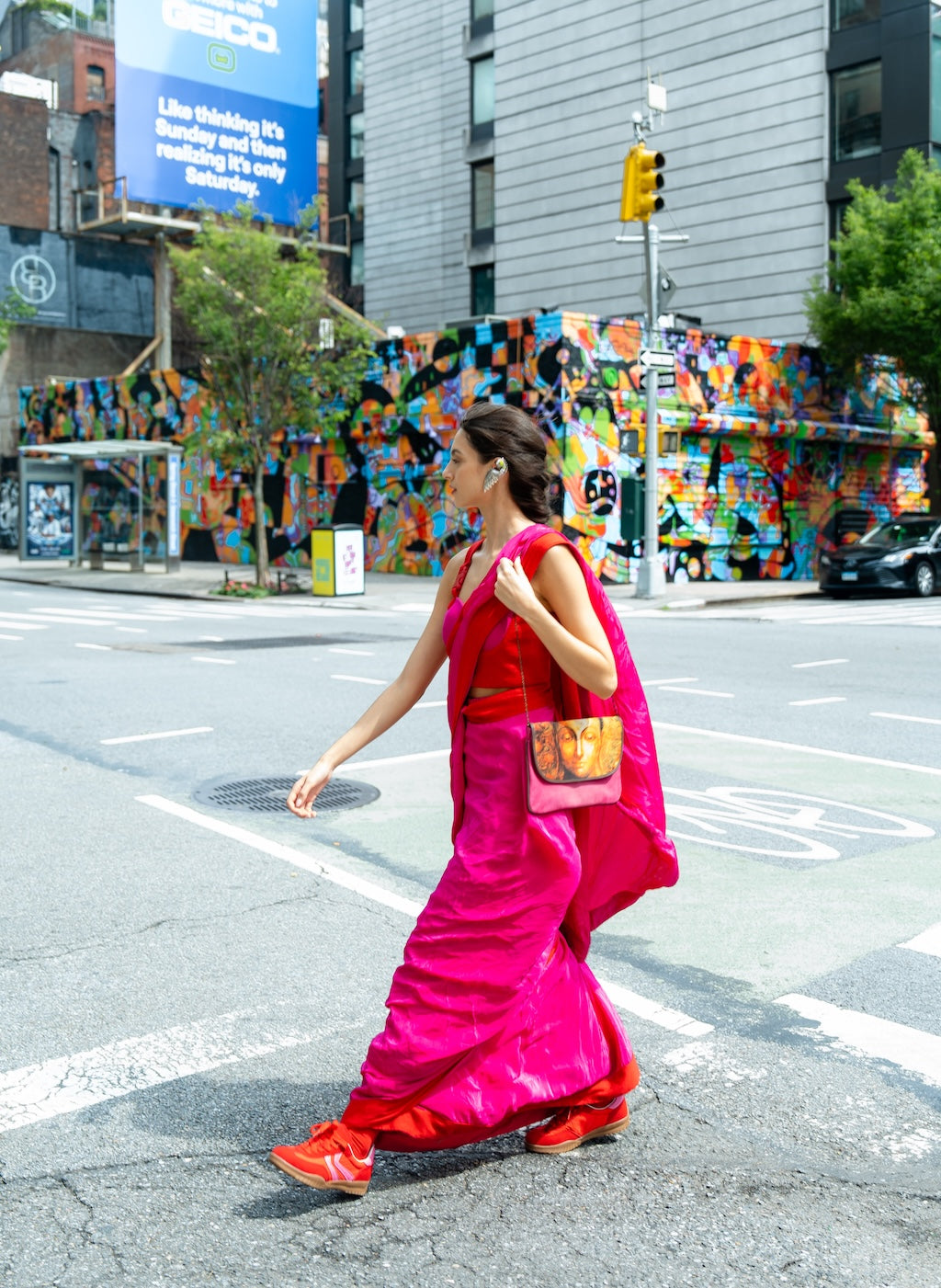 Woman in a bright pink saree (Rebel Roots Buckle Saree) crossing a street with colorful mural in the background. 