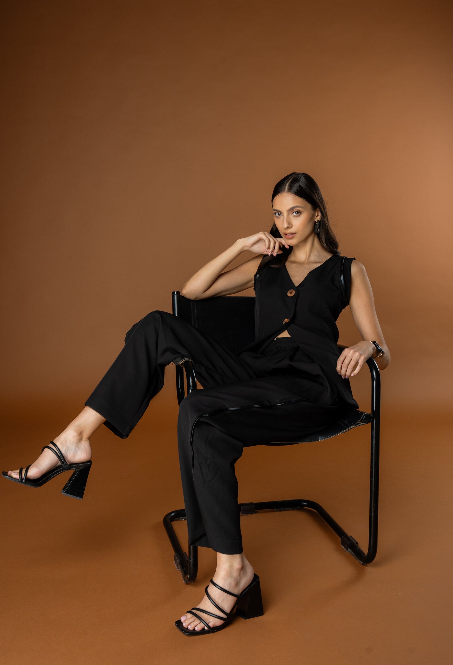 Woman in a black waistcoat and black trouser set from Rebel Roots sitting on a chair against a brown background