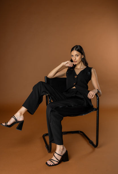 Woman in a black waistcoat and black trouser set from Rebel Roots sitting on a chair against a brown background