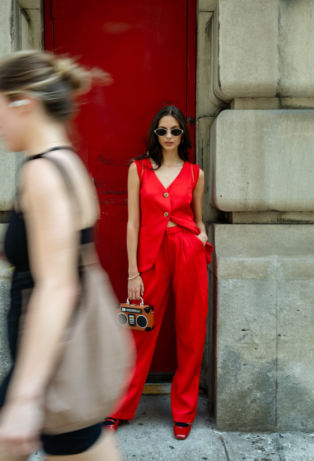 Woman in Rebel Roots Red Capsule PowerSet. Powerset consists of a red waistcoat and Pants. Woman is powerset is standing in front of a red door with a blurred passerby.
