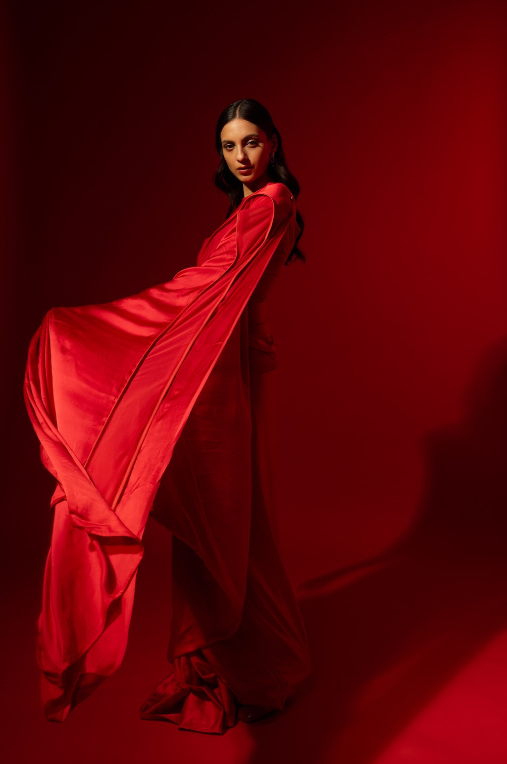 Woman in a flowing red ready to wear Saree standing against a red background. This red Saree is a part of the Rebel Roots Capsule Collection