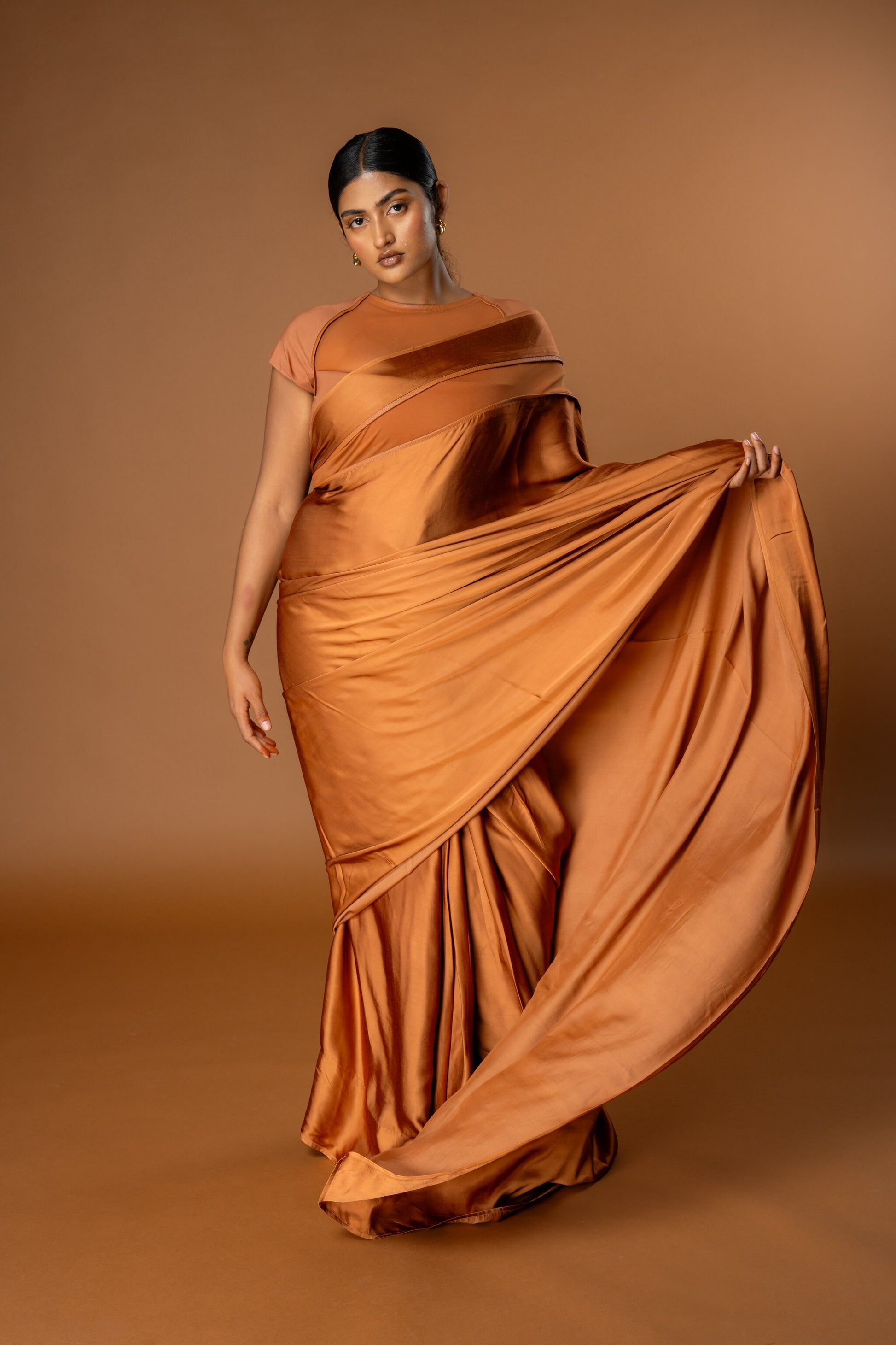 Woman wearing a flowing orange/ Tan Ready to Wear saree against a brown background. The saree is Modern and easy to wear.