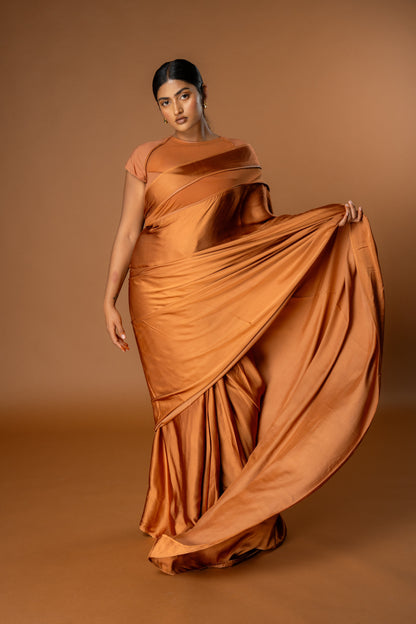 Woman wearing a flowing orange/ Tan Ready to Wear saree against a brown background. The saree is Modern and easy to wear.